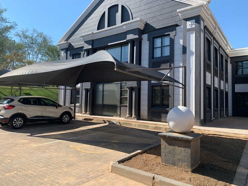 Front exterior view of a modern office building featuring a paved parking area with a shade cloth carport and decorative architectural detailing.