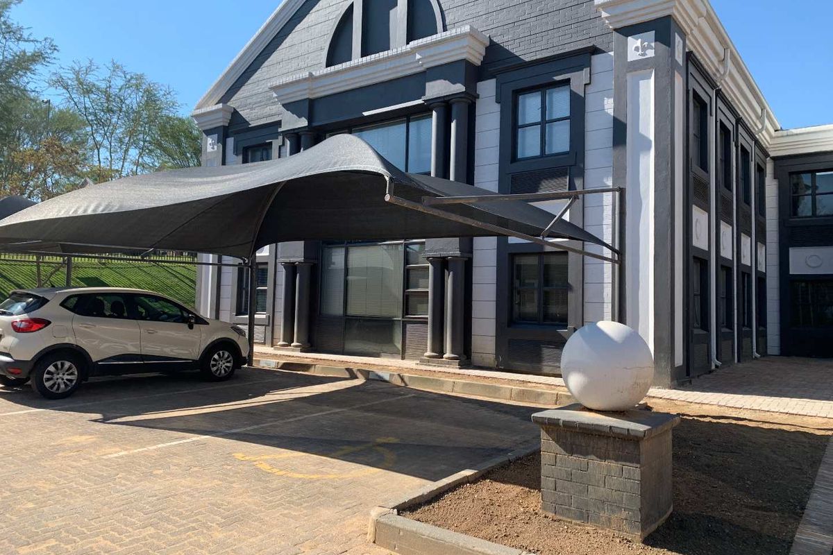 Front exterior view of a modern office building featuring a paved parking area with a shade cloth carport and decorative architectural detailing.
