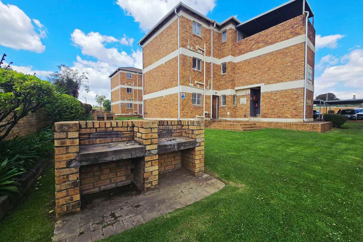 Exterior view of a brick apartment building featuring a communal lawn area and a built-in brick braai facility.