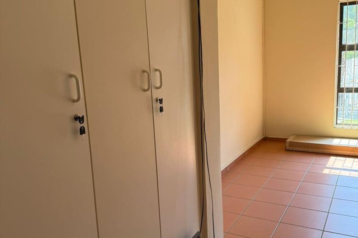 A vacant bedroom featuring built-in cupboards, tiled flooring, and a window with security bars. The room requires minor maintenance to address exposed cabling.