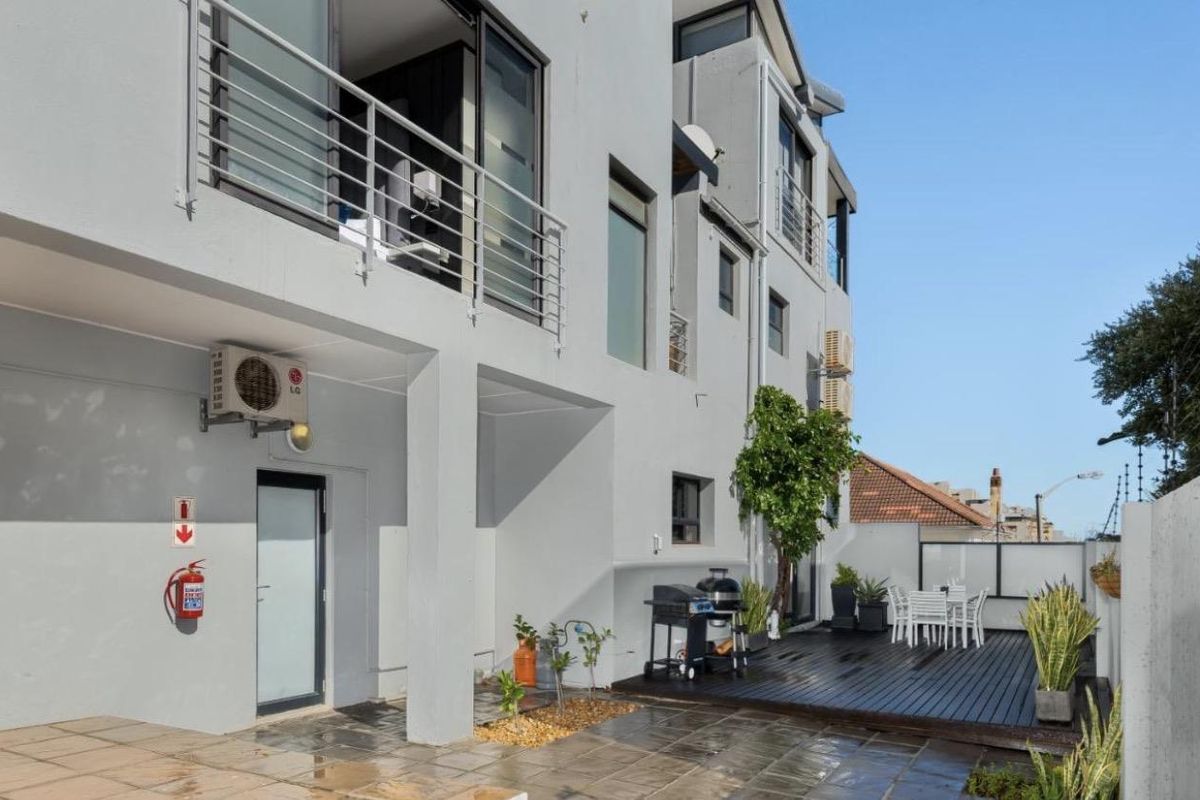 Rear exterior view of the apartment building featuring a paved patio area, timber decking, and upper-level balconies with metal railings.