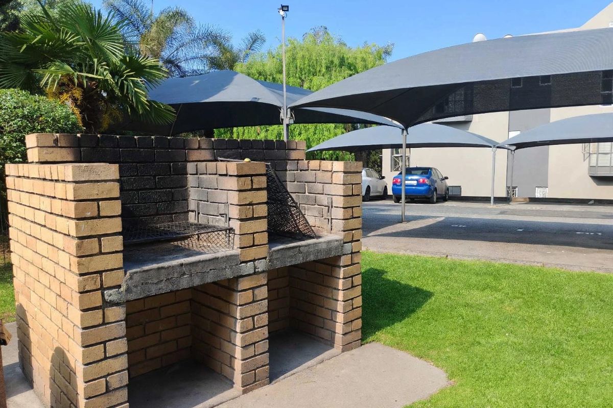 Outdoor communal area featuring a brick built-in braai, paved driveway, and shade sail parking structures set within a landscaped garden.