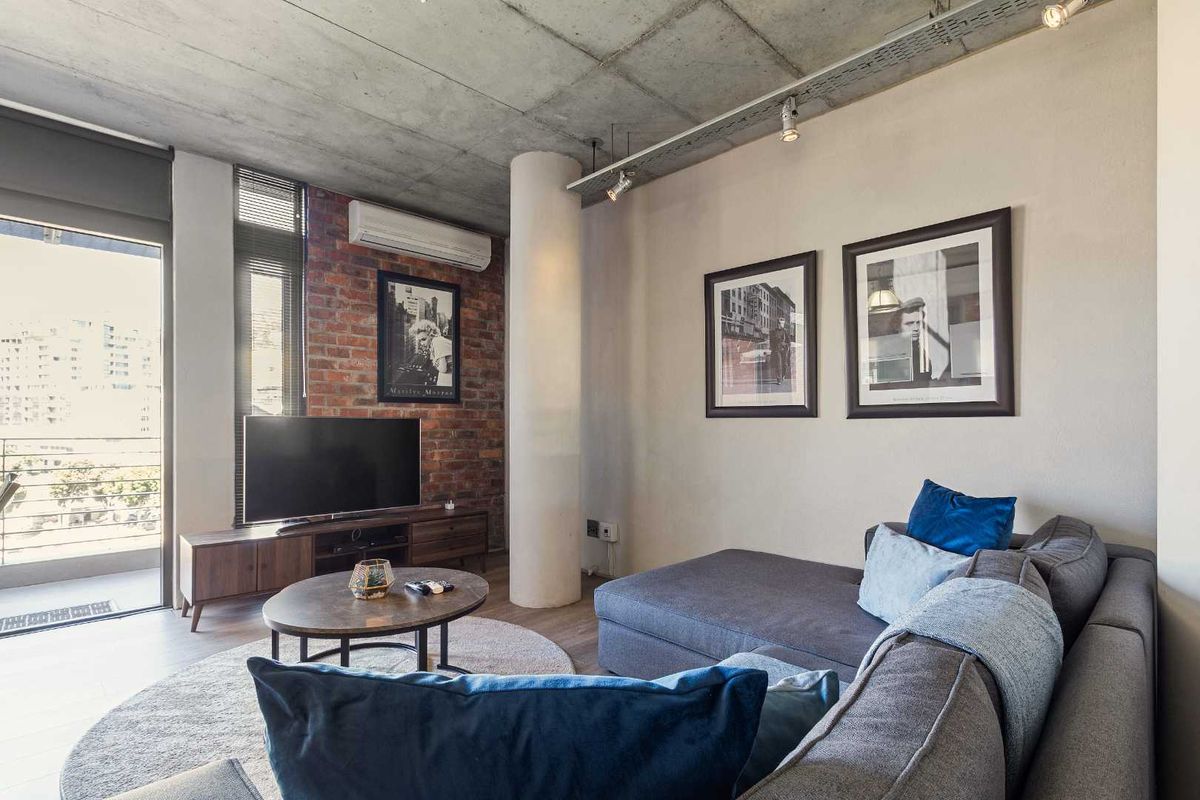 Modern living room featuring an exposed concrete ceiling, industrial brick accent wall, and direct access to a private balcony through sliding glass doors.