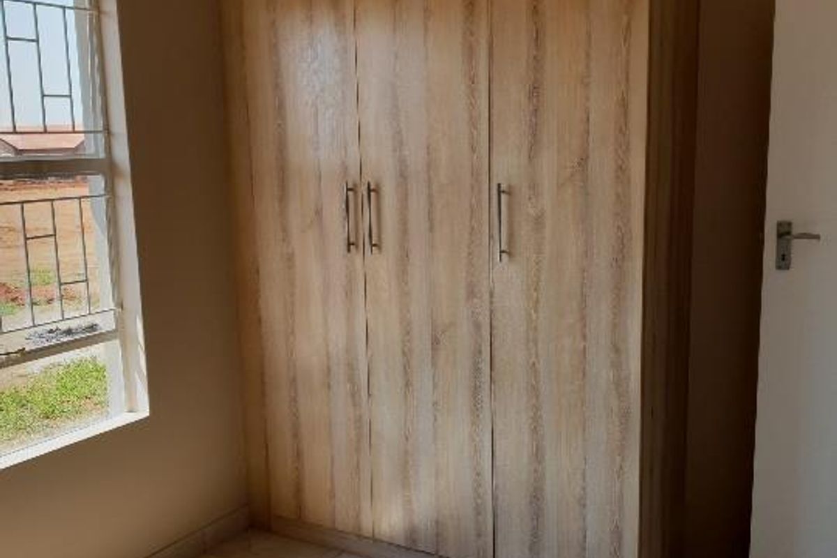 A bright bedroom featuring light wood-finish built-in wardrobes, tiled flooring, and a window with security bars.