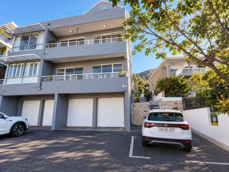 Front elevation of a modern multi-story apartment building featuring multiple garage doors, private balconies with glass railings, and a paved driveway area.
