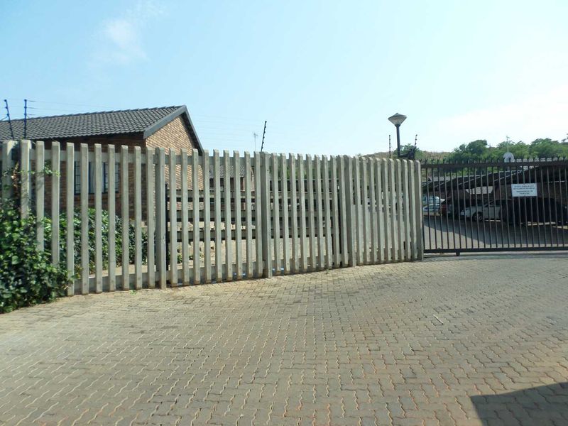Exterior view of the property entrance featuring a paved driveway, concrete palisade fencing, and a secure metal gate leading to a carport area.