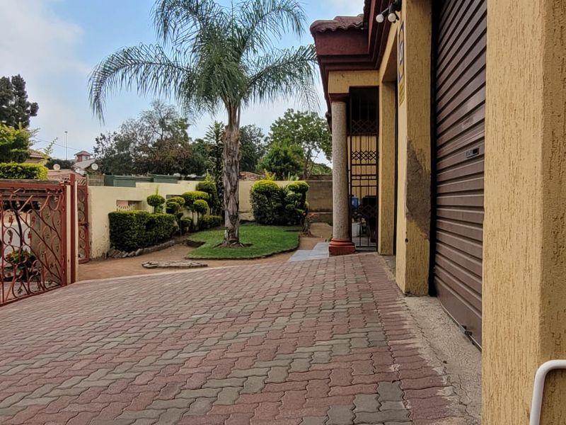 Front exterior view featuring a paved driveway, roller shutter garage door, and a landscaped garden area with a wrought iron entrance gate.