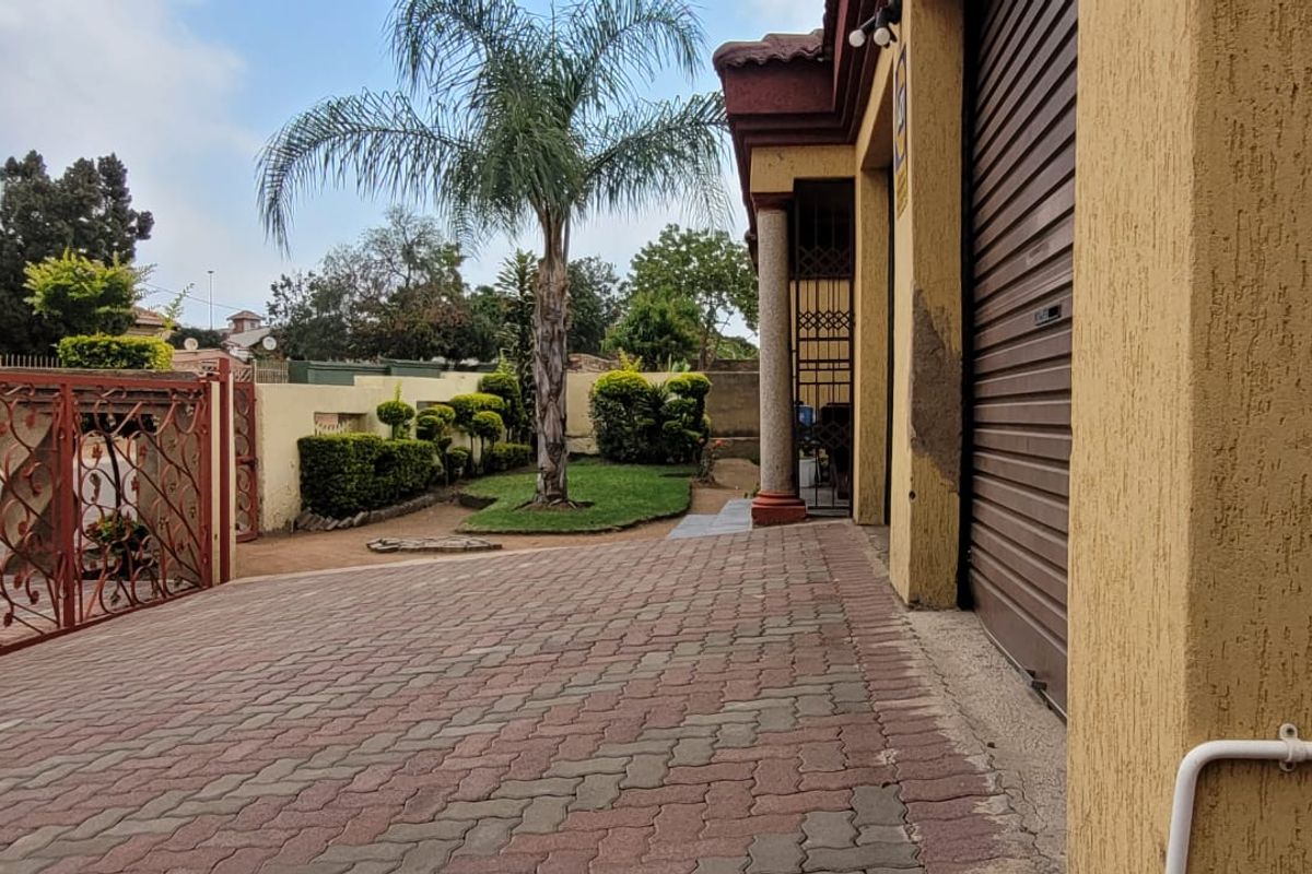 Front exterior view featuring a paved driveway, roller shutter garage door, and a landscaped garden area with a wrought iron entrance gate.