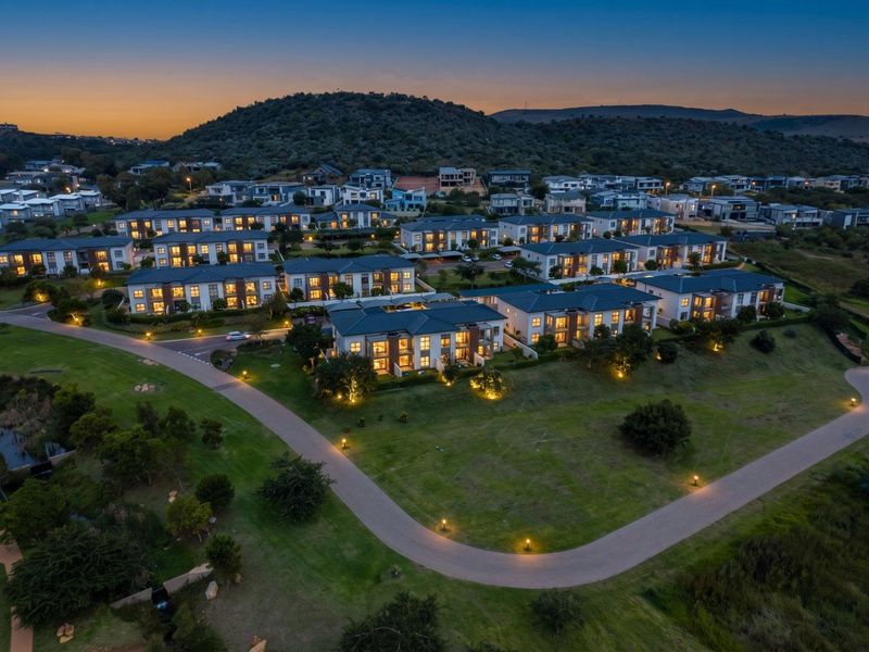 An elevated aerial view of a modern residential apartment complex at dusk, featuring well-maintained landscaping, paved pathways, and exterior building lighting.