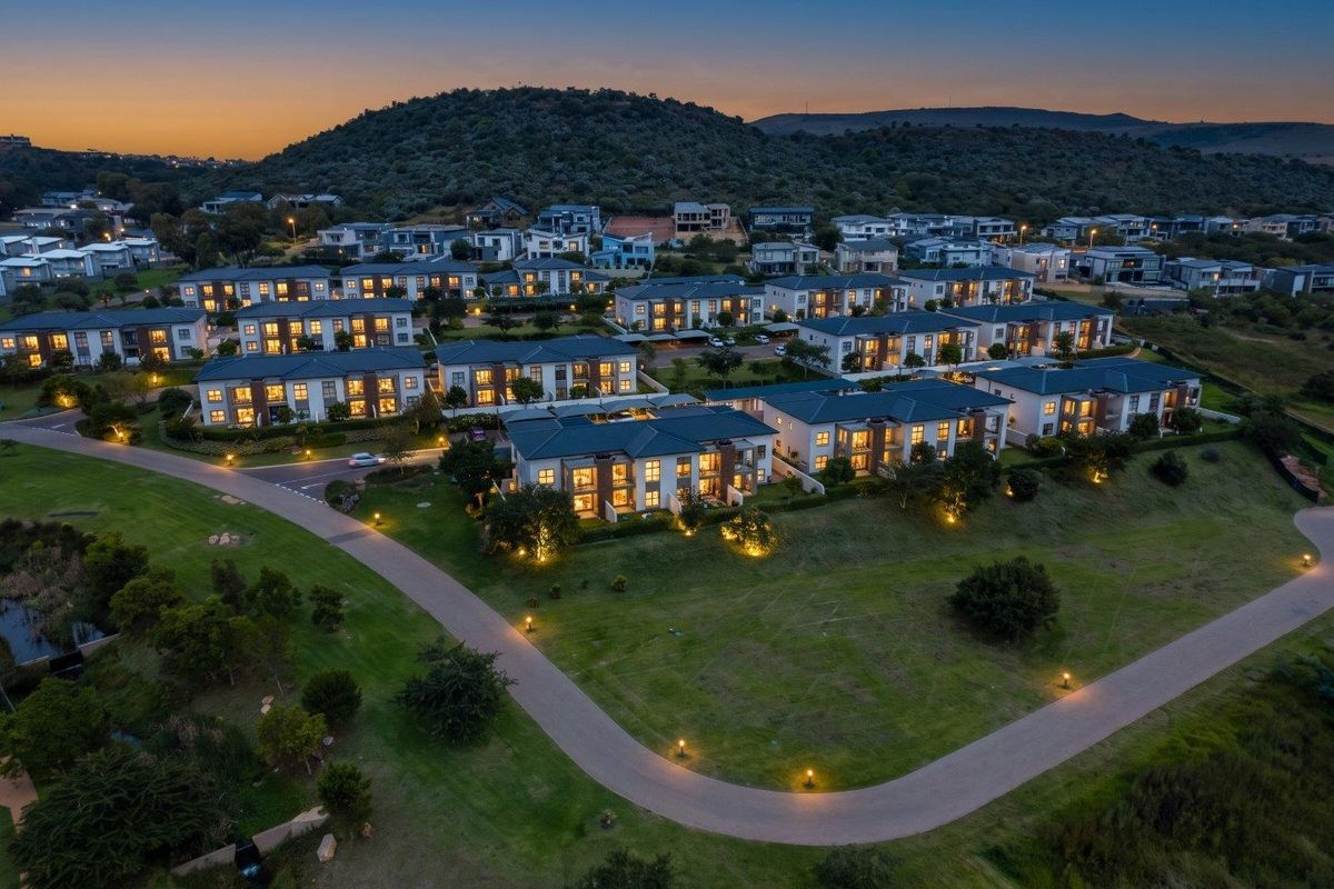 An elevated aerial view of a modern residential apartment complex at dusk, featuring well-maintained landscaping, paved pathways, and exterior building lighting.