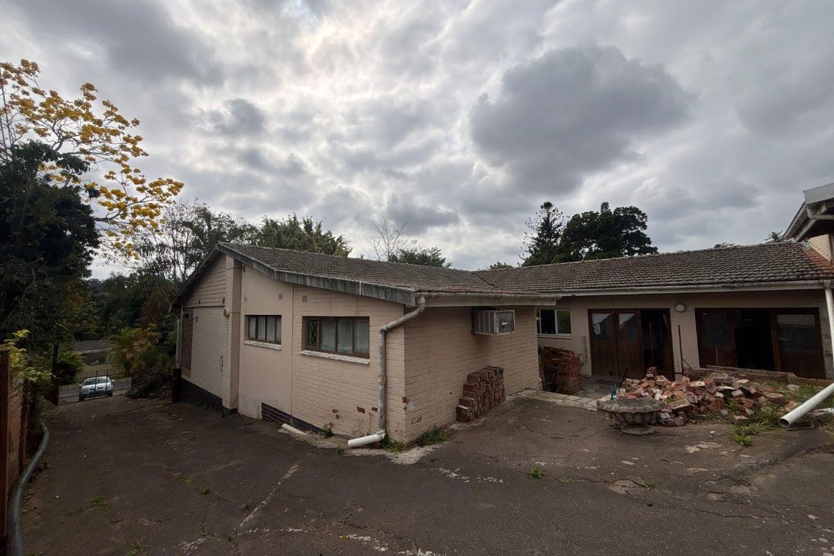 Rear exterior view of a brick house featuring a paved driveway, tiled roof, and visible maintenance requirements including debris removal and exterior repairs.