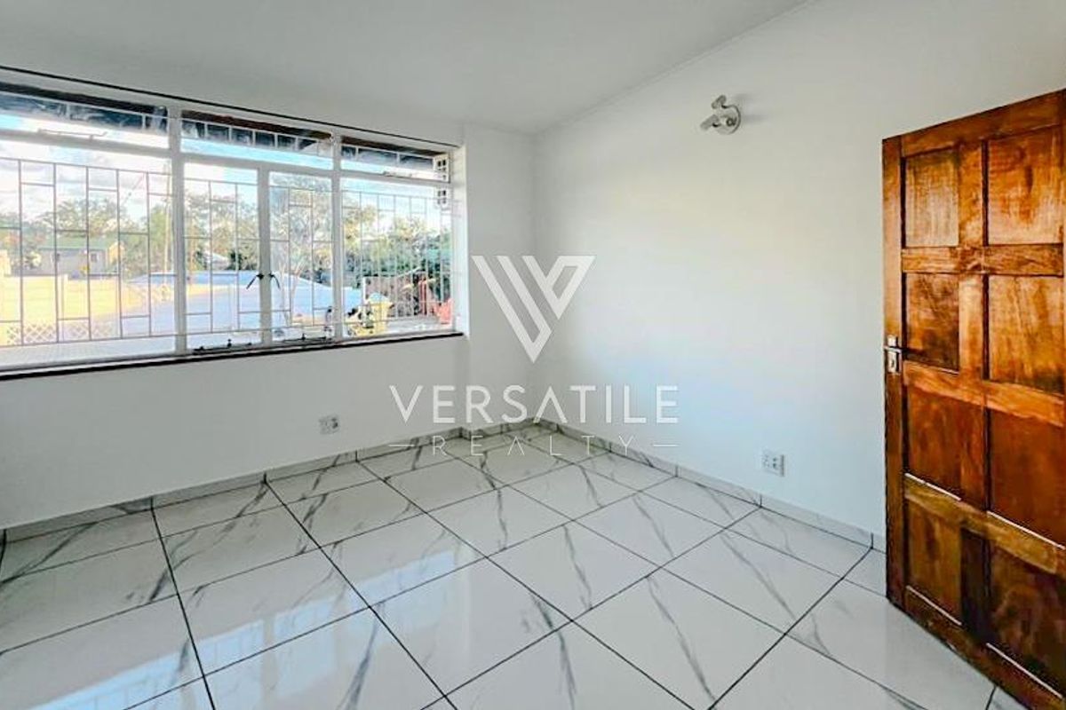 A bright, empty room featuring large windows with security bars, marble-look tiled flooring, and a traditional wooden door.