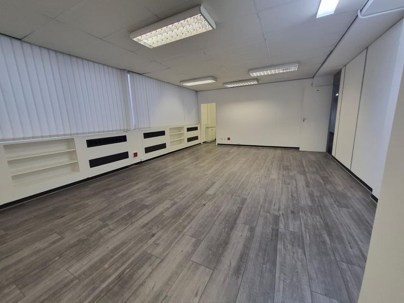 A spacious, empty office area featuring modern laminate flooring, built-in shelving units, vertical blinds, and a suspended ceiling with integrated lighting.