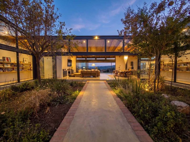 A stunning modern residence featuring expansive glass walls, exposed structural beams, and a beautifully landscaped central courtyard pathway at dusk.