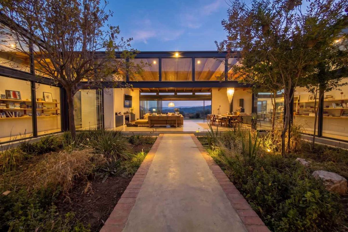 A stunning modern residence featuring expansive glass walls, exposed structural beams, and a beautifully landscaped central courtyard pathway at dusk.