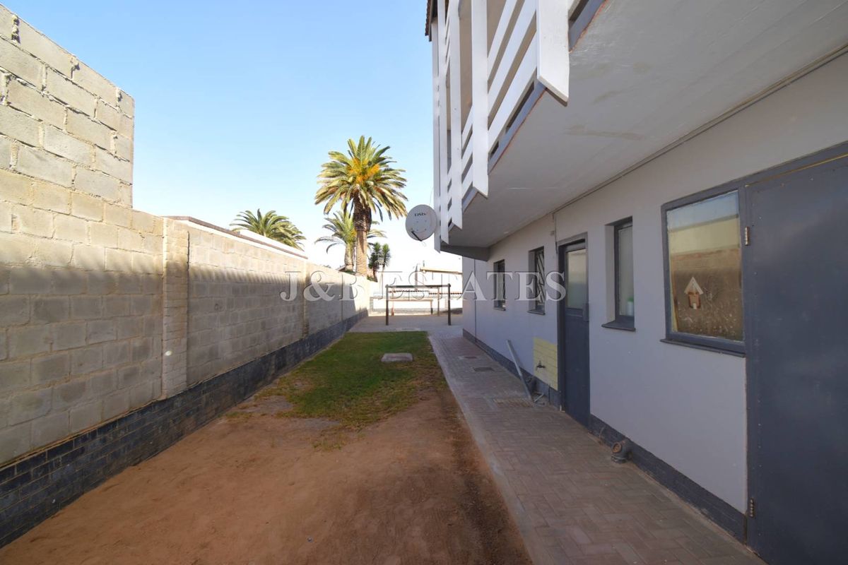 A view of the rear exterior area of the apartment, featuring a paved walkway, a small patch of lawn, and a boundary wall.