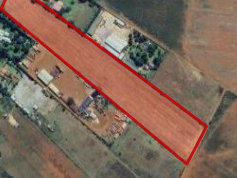 An aerial view of a vacant agricultural land plot, clearly outlined in red, showing the surrounding terrain and nearby structures.