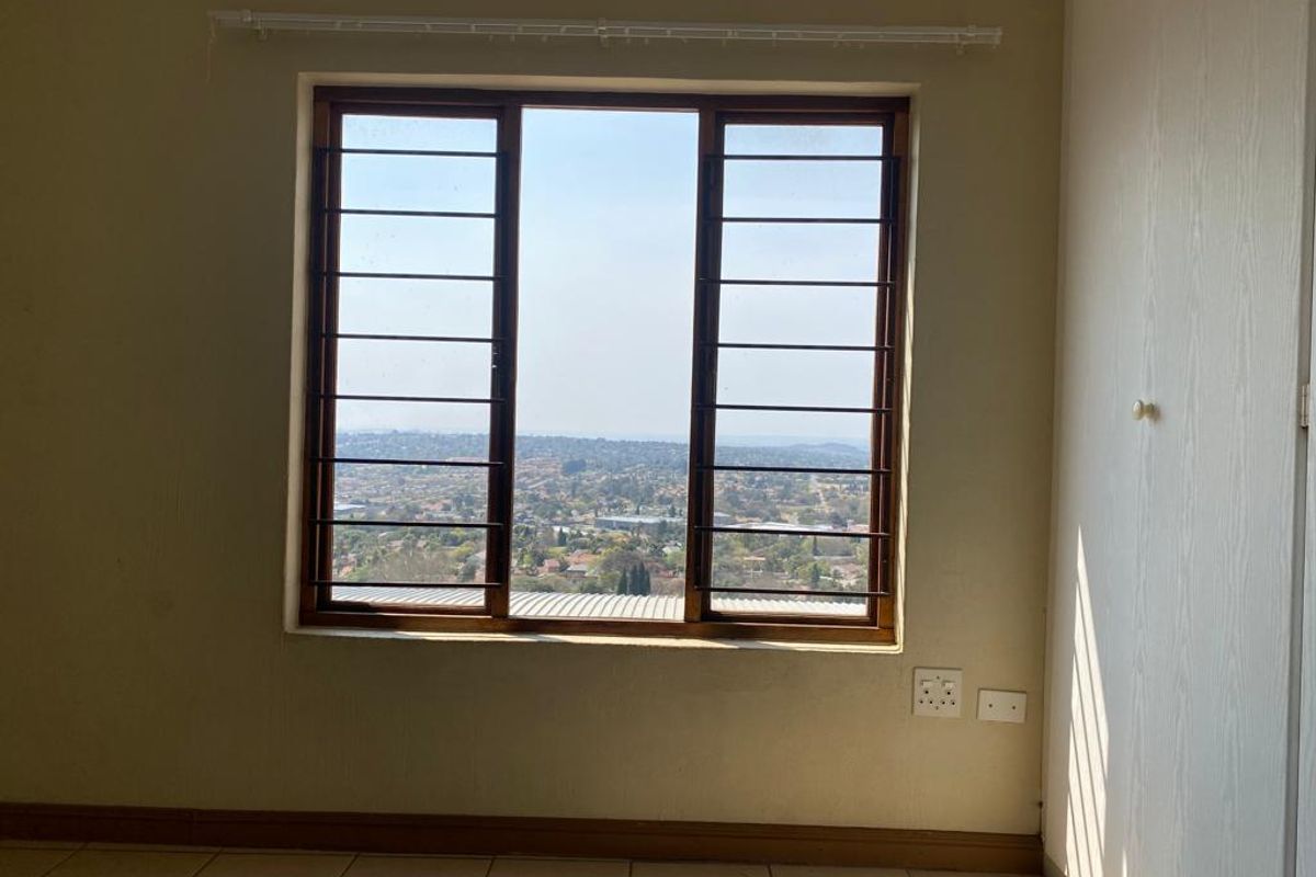 A bright bedroom featuring built-in cupboards, tiled flooring, and a large window with security bars offering expansive elevated views.