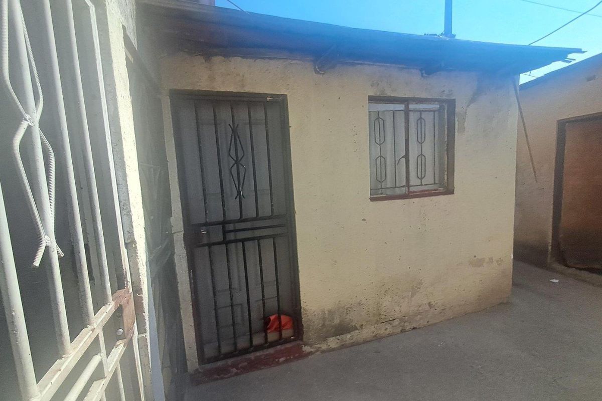 Exterior view of a small residential structure featuring a security gate and window bars, showing signs of significant wear and weathering.