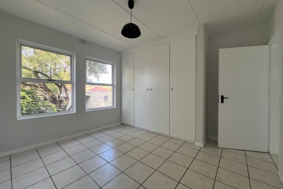 A bright, empty bedroom featuring neutral tiled flooring, built-in cupboards, and large windows providing natural light and a pleasant exterior view.