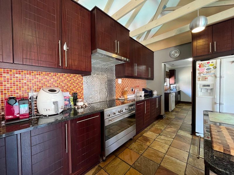 A spacious kitchen featuring dark wood cabinetry, granite countertops, a mosaic tile backsplash, and exposed timber roof trusses, with an adjoining scullery area.