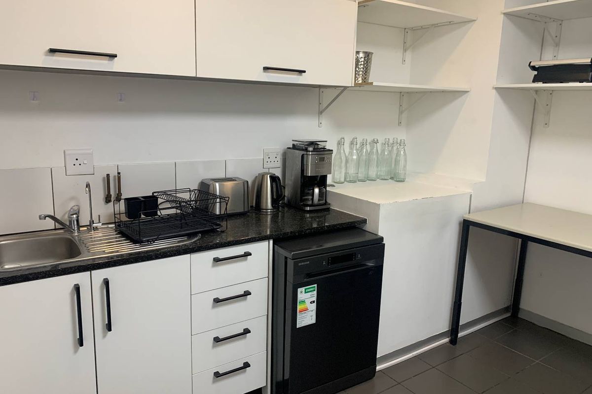 A functional office kitchenette featuring white cabinetry, a stainless steel sink, dark laminate countertops, and a built-in dishwasher.
