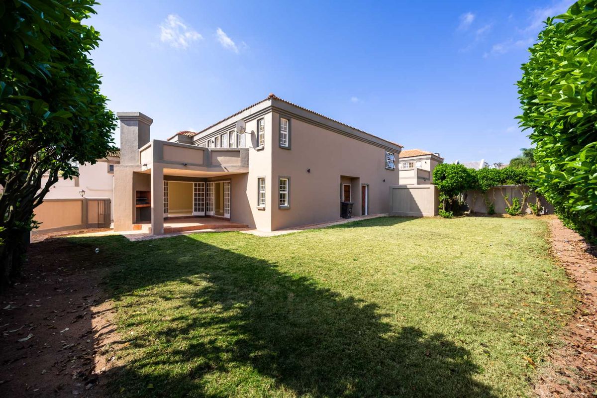 Rear view of the property featuring a spacious lawn, a covered patio with a built-in braai, and a well-maintained exterior facade.