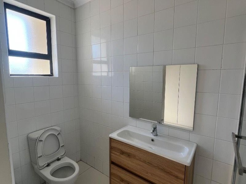 A clean, modern bathroom featuring a wall-mounted wood-finish vanity, ceramic basin, toilet, and full-height white wall tiling.