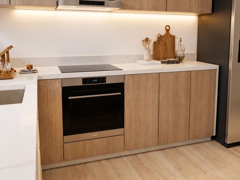 Modern kitchen featuring light wood cabinetry, stone countertops, integrated oven, induction cooktop, and stainless steel extractor fan with under-cabinet lighting.