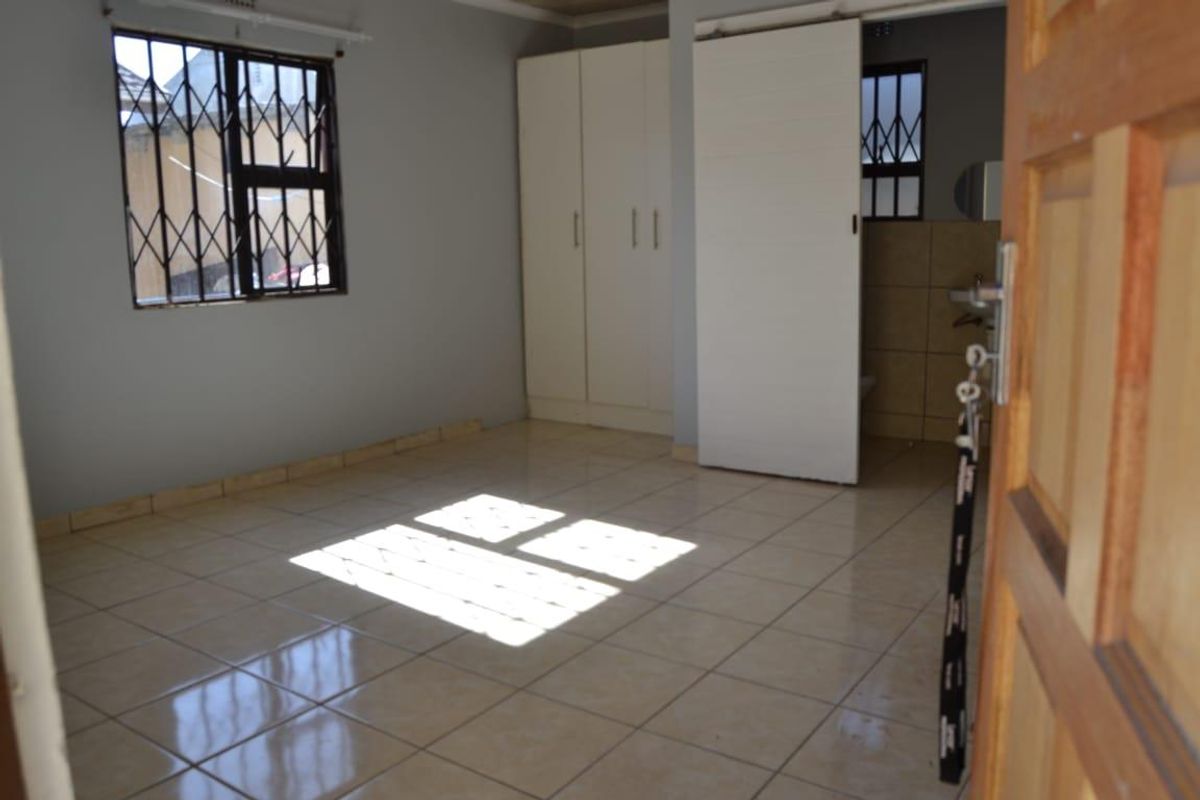 A bright, empty bedroom featuring tiled flooring, built-in cupboards, and security bars on the window, with an adjacent en-suite bathroom area.