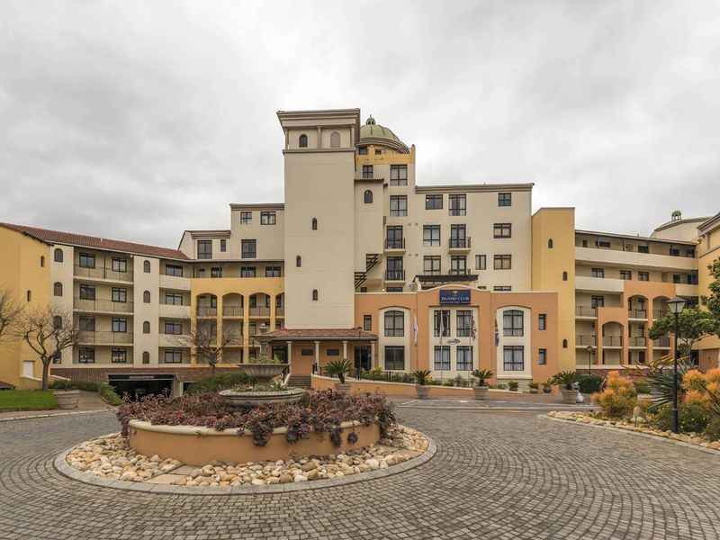 Front elevation of a multi-story apartment complex featuring a paved circular driveway, central fountain, and landscaped garden beds.