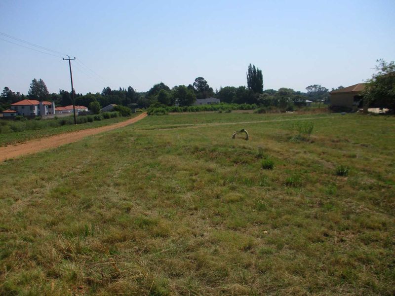 A vacant residential plot of land featuring open grass, a dirt access road, and nearby utility infrastructure in a quiet neighborhood setting.