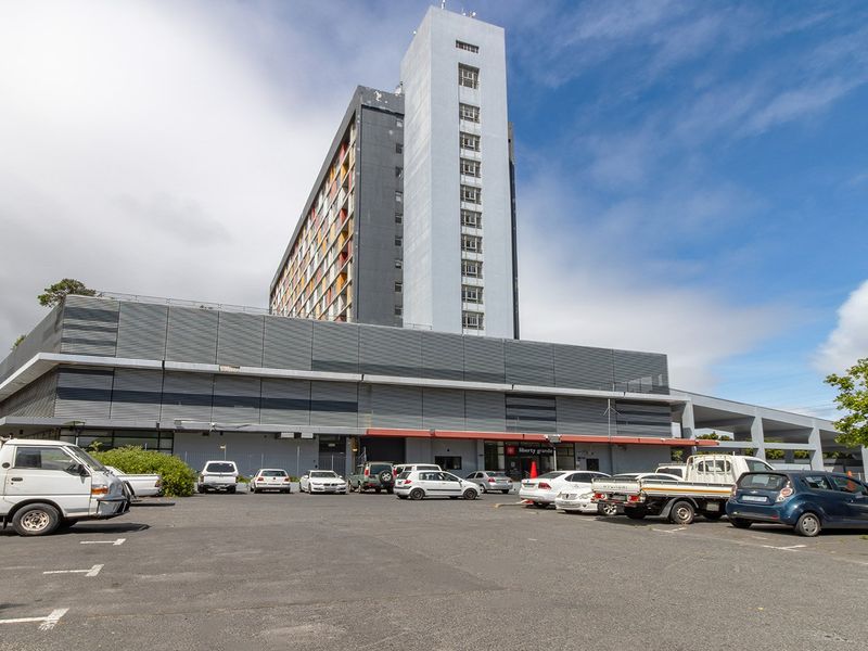 Exterior view of a multi-story apartment building featuring a large asphalt parking area and metal-clad facade.