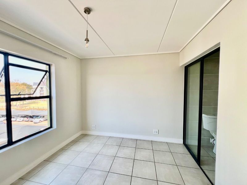 A bright, empty bedroom featuring neutral tiled flooring, modern aluminium window frames, and an en-suite bathroom accessible via a sliding glass door.