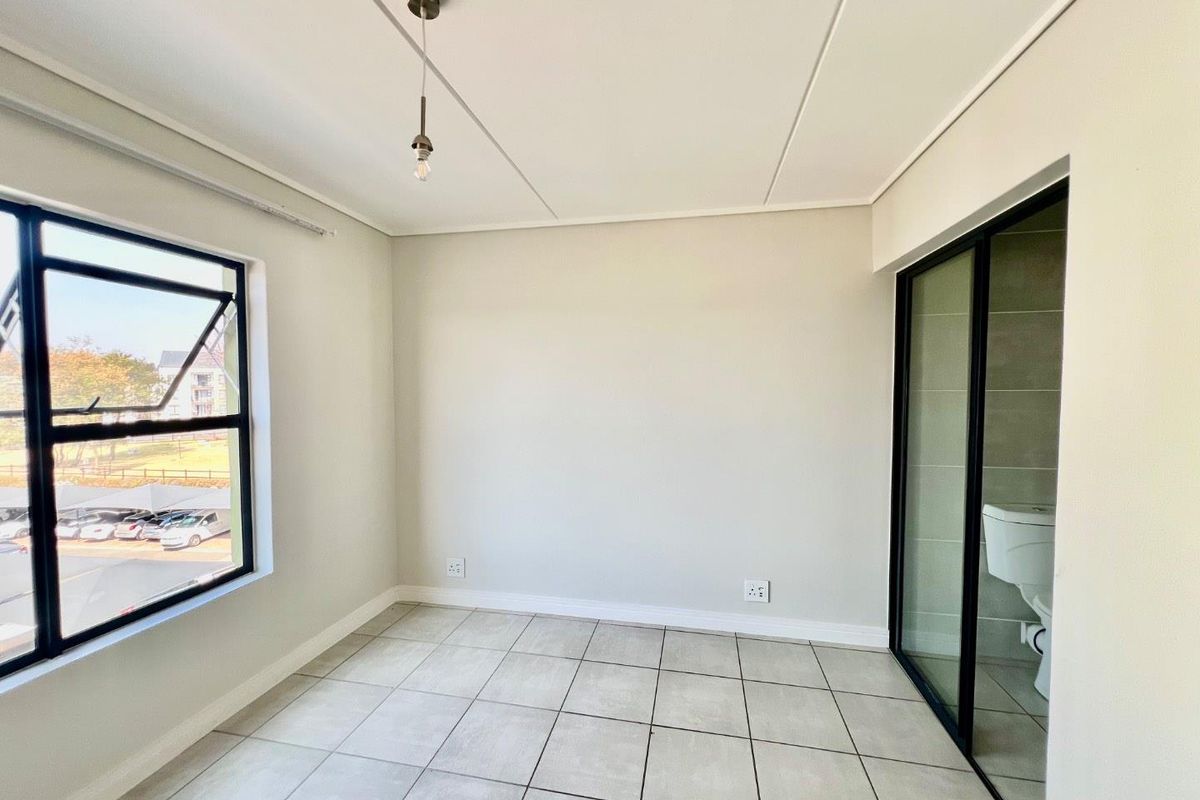 A bright, empty bedroom featuring neutral tiled flooring, modern aluminium window frames, and an en-suite bathroom accessible via a sliding glass door.