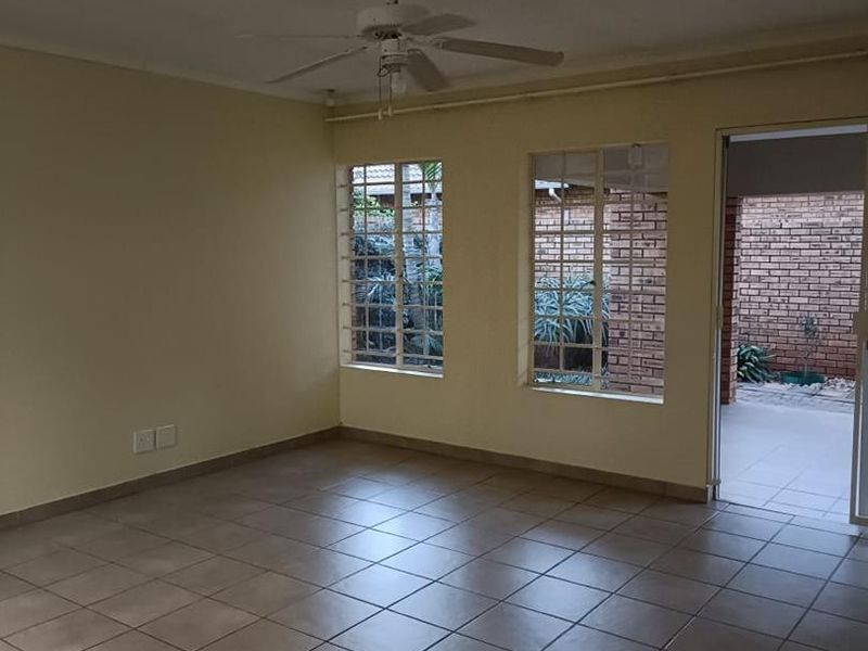 An empty living room featuring tiled flooring, a ceiling fan, and large windows with security bars, providing access to an outdoor area.