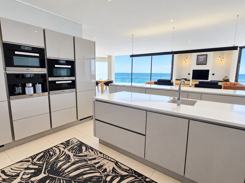 Modern open-plan kitchen featuring high-end integrated appliances, sleek cabinetry, and a large island, seamlessly flowing into the living area with panoramic ocean views.