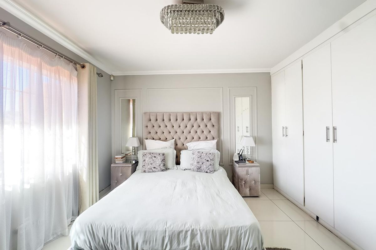 A well-maintained bedroom featuring built-in cupboards, tiled flooring, decorative wall molding with integrated mirrors, and a central crystal chandelier.