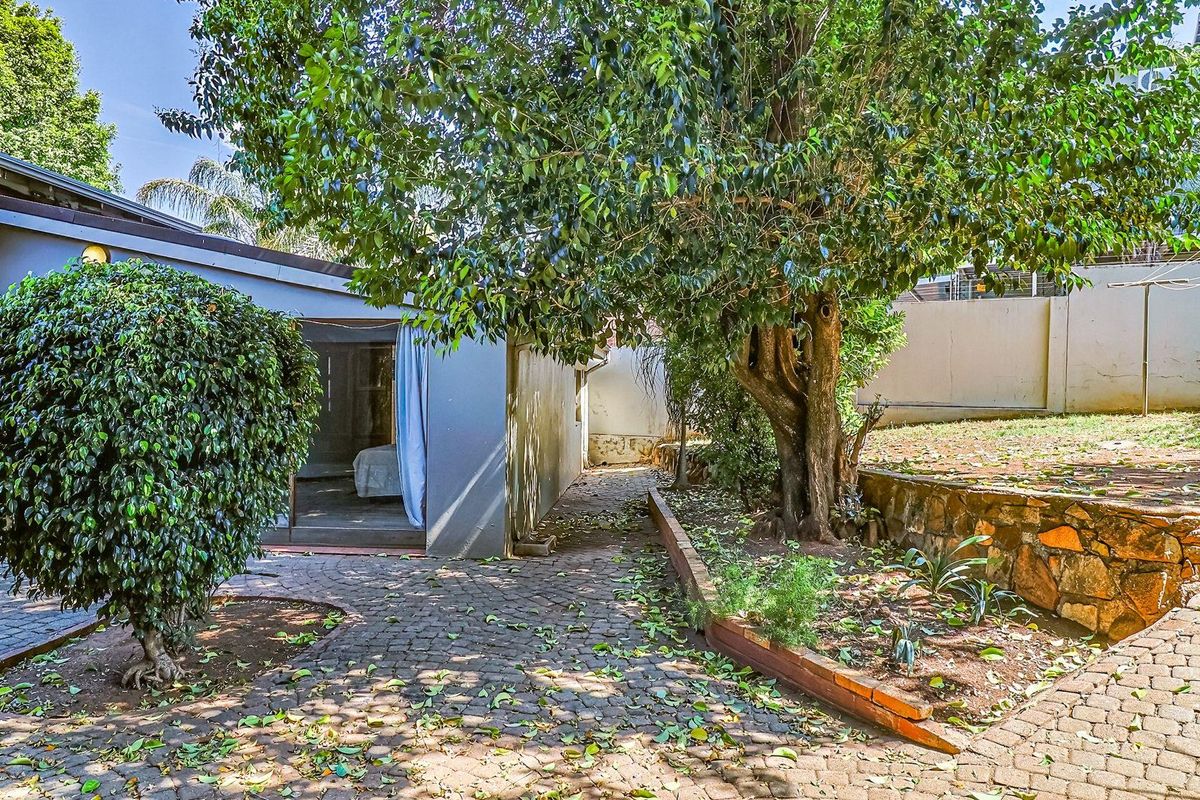 A rear garden area featuring paved pathways, a stone retaining wall, and mature trees, providing access to the main dwelling.