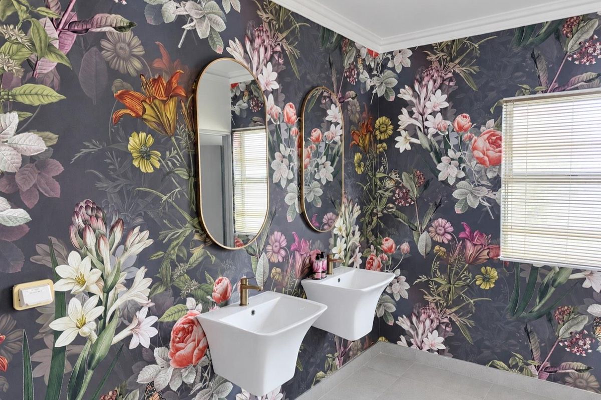 A modern bathroom featuring dual wall-mounted basins with brass fixtures, set against a bold floral wallpaper and complemented by neutral floor tiles.