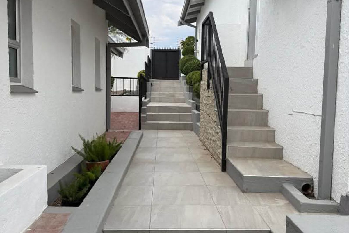 A well-maintained exterior walkway featuring tiled steps, metal handrails, and stone cladding accents, leading towards a secure rear gate.