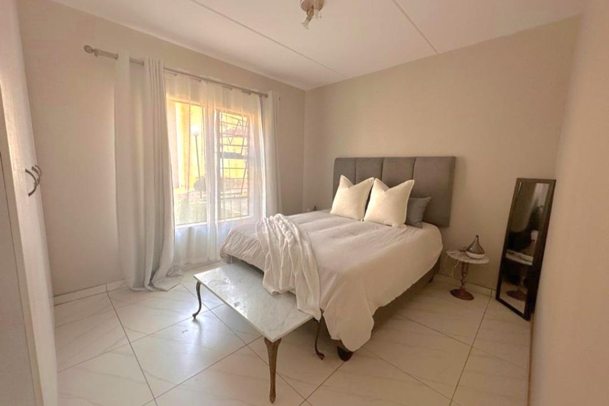 A bright bedroom featuring neutral tiled flooring, a large window with security bars, and a simple ceiling light fixture.