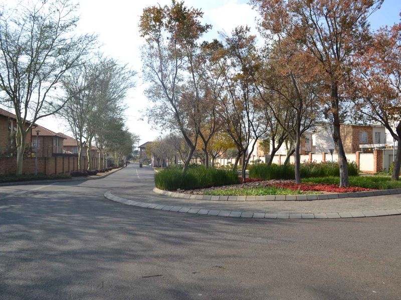 A view of the residential street approach featuring paved roads, mature trees, and landscaped garden beds within the estate.