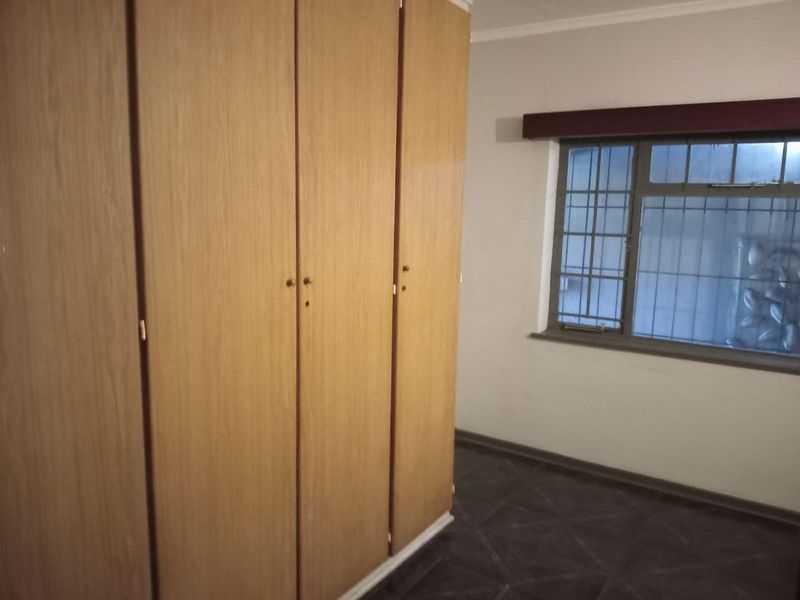 A bedroom featuring built-in wooden cupboards, tiled flooring, and a window fitted with security bars.