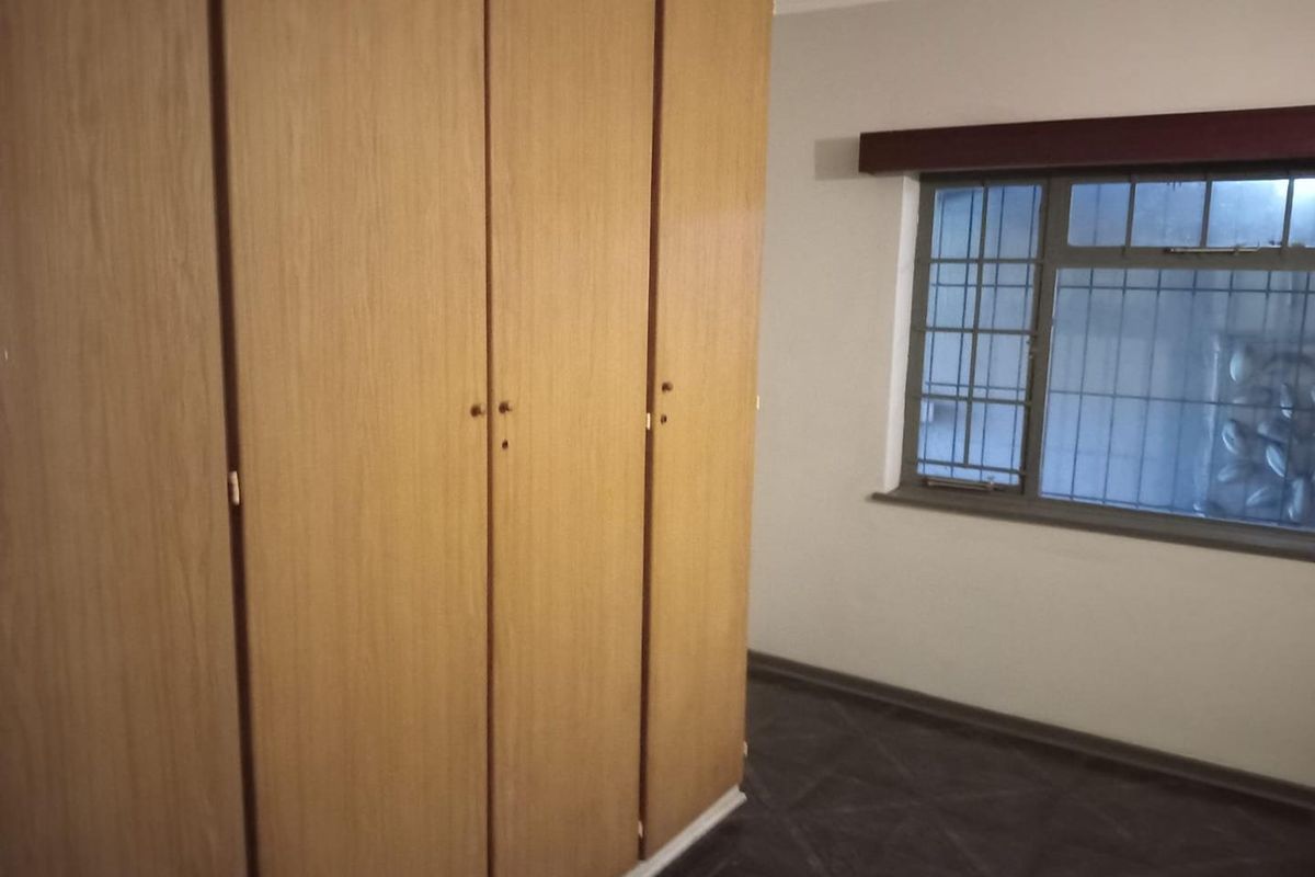 A bedroom featuring built-in wooden cupboards, tiled flooring, and a window fitted with security bars.