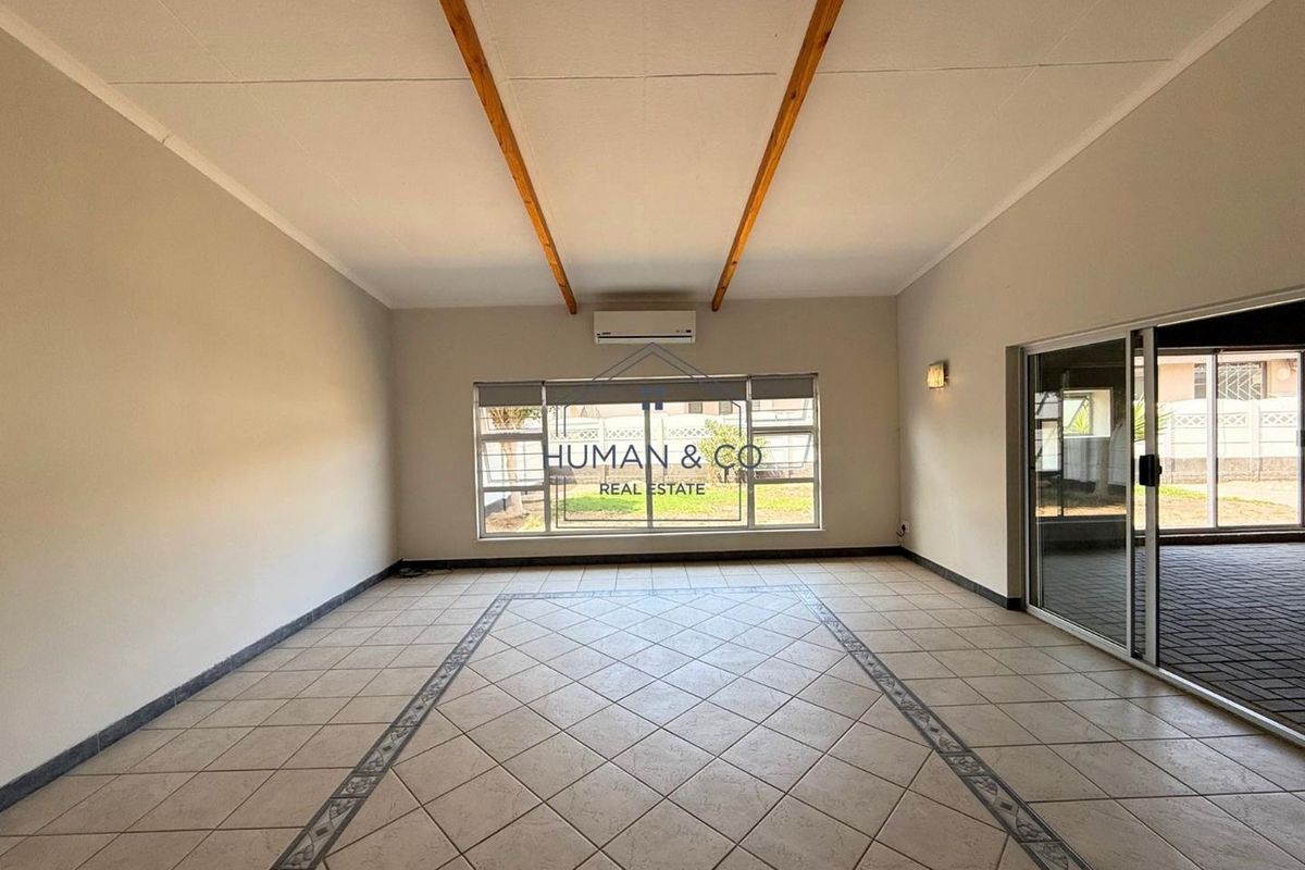 Spacious living area featuring tiled flooring, exposed timber ceiling beams, a wall-mounted air conditioning unit, and large sliding glass doors leading to the exterior.