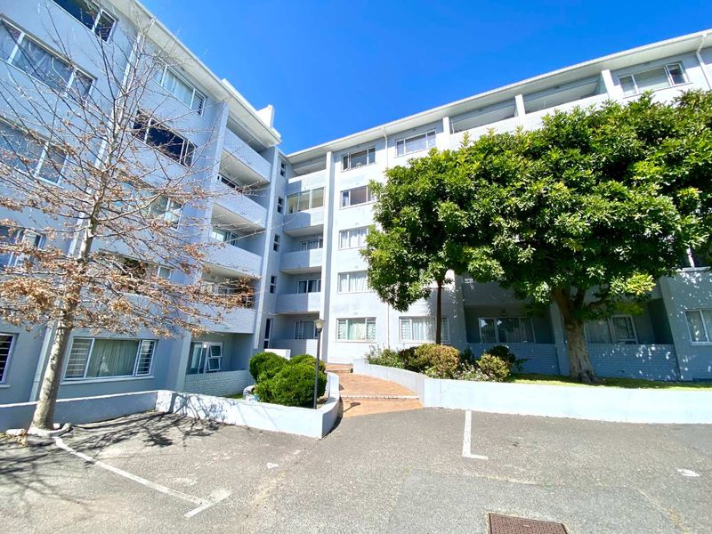 Exterior view of a multi-story apartment building featuring paved walkways, balconies, and an asphalt parking area.