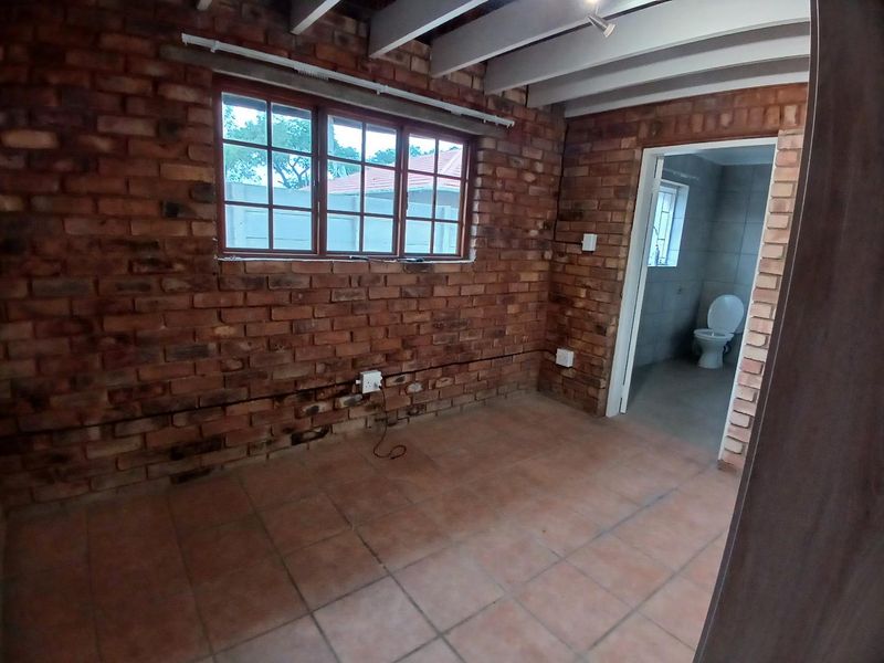 An empty bedroom featuring rustic exposed brick walls, exposed timber ceiling beams, and tiled flooring, with direct access to an en-suite bathroom.