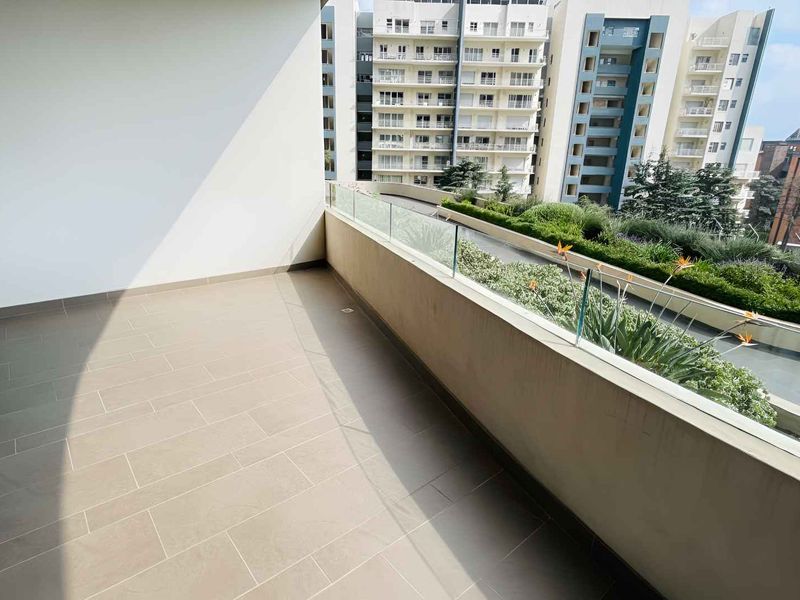 A spacious, modern balcony featuring clean tiled flooring and a sleek glass balustrade, offering an open view of the surrounding urban landscape.