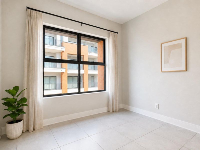 A bright, empty bedroom featuring neutral tiled flooring, clean walls, and a large black-framed window overlooking the apartment complex.